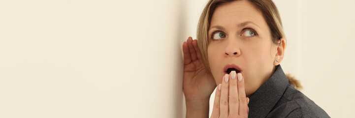 Curious woman puts ear close to beige wall covering mouth