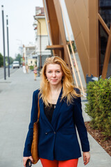 young smiling female woman on city street. 39 year old white caucasian woman with long honey color hair, dark blue jacket, red jeans, casual clothes. three quarter length view portrait, vertical photo