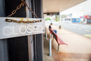 “Sorry, we are closed” sign hanging on shop door. Unrecognizable person sitting on the bench outside.