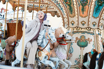 Obraz na płótnie Canvas Cheerful woman in a faux fur coat and mittens rides a carousel and has fun