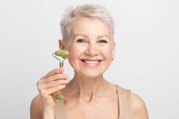 Obraz na płótnie Canvas Elderly woman with short hair massaging face skin using natural facial stone trendy jade green quartz roller