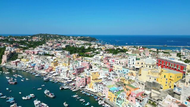 Enthused aerial pullback from Panoramica sulla Corricella on Procida Island (Isola di Procida) of the cobalt Tyrrhenian Sea in the Gulf of Naples with pastel buildings, white boats, wakes, breakwaters