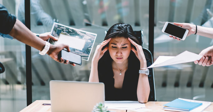 Young disappointed woman feeling headache from deadline surrounded by colleagues at workplace