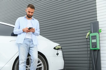 Beautiful young stylish man is with electric car at daytime charging the vehicle