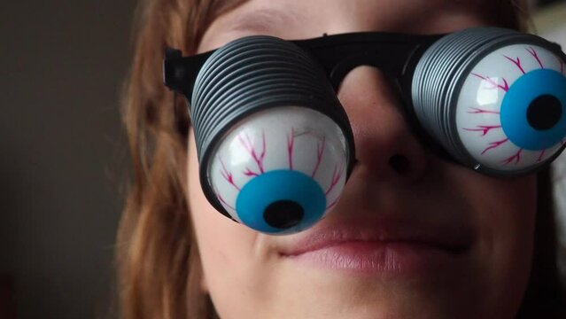 A 9-year-old caucasian girl wearing glasses with eyeballs falling out on springs. A child is preparing for Halloween and trying on funny, scary accessories. Masquerade decoration. Black humor