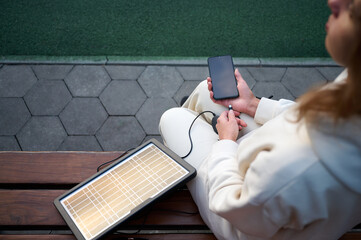Pretty woman sitting on the bench and charging her phone with a portable solar panel. Solar battery...
