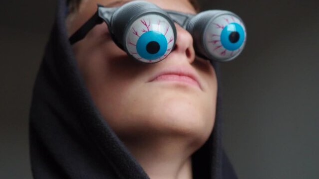 A 10-year-old Caucasian boy wearing glasses with eyeballs falling out on springs. A child is preparing for Halloween and trying on funny, scary accessories. Masquerade decoration. Black humor