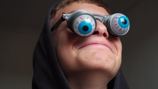 A 10-year-old Caucasian boy wearing glasses with eyeballs falling out on springs. A child is preparing for Halloween and trying on funny, scary accessories. Masquerade decoration. Black humor.