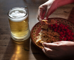 Concept of handling food, hands taking corn tortilla chips and spicy peanuts from a Mexican clay plate, on a wooden table with a jar of cold beer.