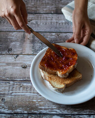 Concept of food handling, grabbing or taking food with hands. female hands spreading jam on a slice of bread on a white plate on a rustic wooden table, with a napkin on the side, top view flat lay.