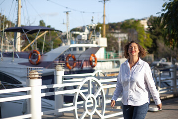 Beautiful fat woman walking on the city street against the yacht. The plus size model dressed white t-shirt, jeans and red lips. The basic outfit on the large model looks stylish