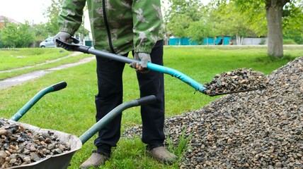 a man in work clothes loads gravel with a shovel into a wheelbarrow on a green lawn in the...