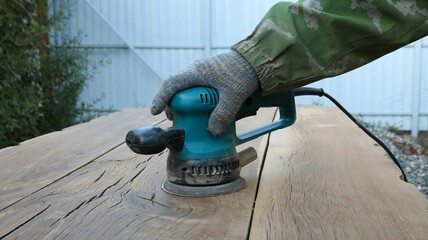 carpenter in gloves works with a sander in a green case with a round emery attachment on polishing solid wooden oak boards in an outdoor workshop, smoothing the surface of the wood with emery - obrazy, fototapety, plakaty