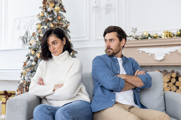 Frustrated and upset family couple sitting on sofa in living room on christmas, man and woman quarreling not talking, celebrating new year and christmas near tree on winter day