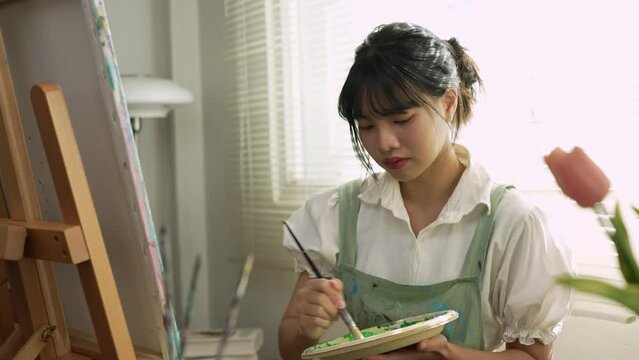 Young Asian female artist holds a palette with mixed acrylic paints. Use your creativity in designing pictures. Create art on canvas Develop art drawing skills Hobby activities in the studio or home.