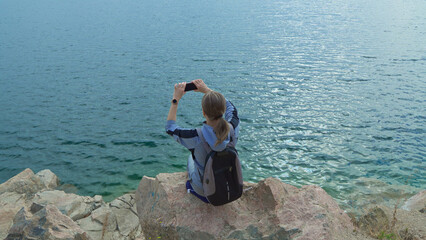 Young female tourist sits on mountainside above ocean and takes pictures of seascape on her smartphone.