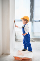 Child construction worker painting wall with paint brush in apartment. Kid in safety helmet and work overalls using paint brush while working at home under renovation.