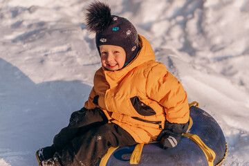 Fototapeta na wymiar Active toddler boy in a yellow jacket sliding down the hill on snow tube.Winter fun,active lifestyle concept.