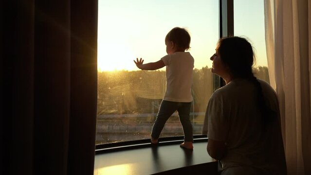 Perched on windowsill toddler girl under supervision of mother looks through window at sunset. Colors of sun give tenderness to picture