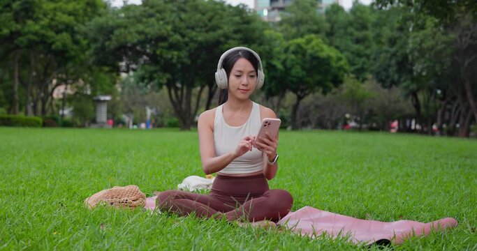 Woman do yoga and sit on the mat, wear the headphone use of mobile phone at park