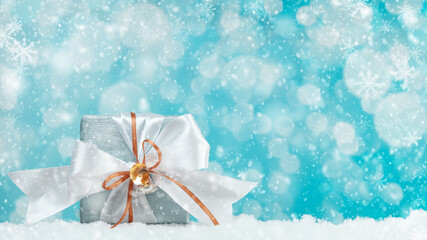 Gift box wrapped in blue silver paper with white and gold ribbon bow. Blue background, snow and bokeh light. Christmas and New Year gifts, Boxing Day.