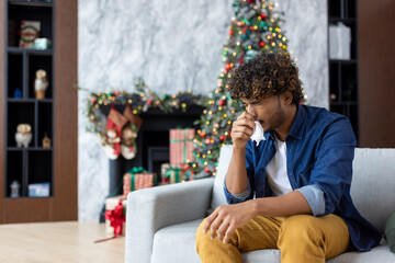 Sick sneezing man with runny nose sitting on sofa at home, hispanic winter day near Christmas tree, cold and allergy in living room inside house.