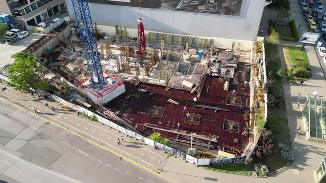 Top-down aerial view of an active urban construction site with a tower crane, concrete pump, formwork, reinforcement and workers building the structural foundation and lower floors of a new developmen