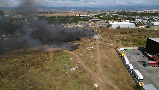 Aerial footage of a fire burning and spreading along a garbage field next to the largest residential neighbourhood of Bulgaria. Lyulin illegal dump on fire. Firefighters putting out fire. Drone shot.
