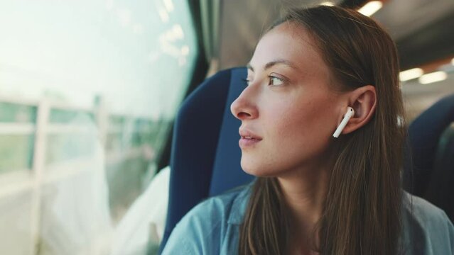 Close up, cute girl with long brown hair, wearing blue shirt, wearing wireless earphone into ear traveling by suburban train