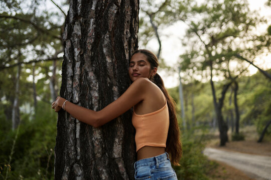 Teenage girl with eyes closed hugging tree trunk in forest at sunset
