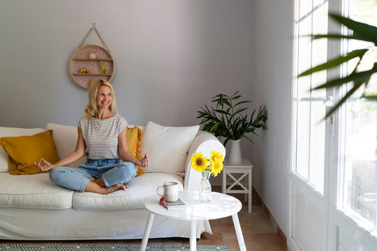 Smiling mature woman meditating on sofa in living room at home