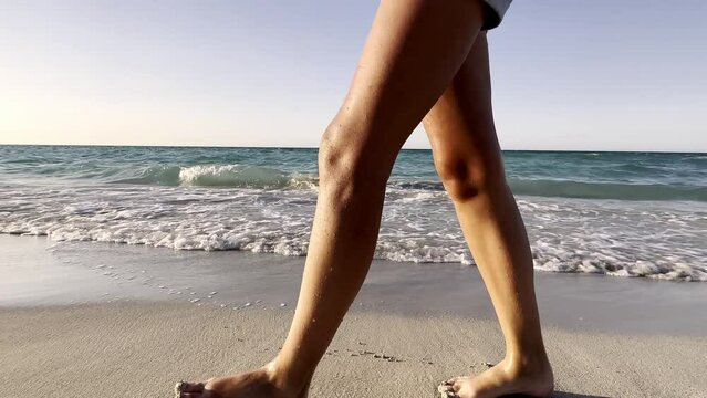 Women's feet of tourists walking barefoot along the shore at sunset. Legs of a young woman walking along the ocean beach during sunrise. The girl steps on the wet sand of the coastline. Slow motion