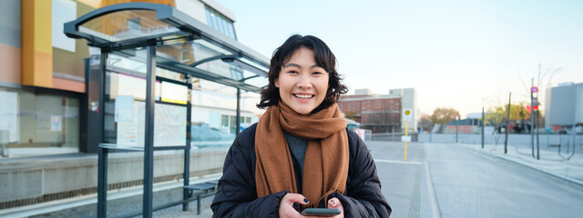 Portrait of Korean woman in winter jacket, standing with smartphone, waiting for bus on stop,...