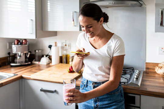 Happy woman photographing milkshake through smart phone in kitchen at home