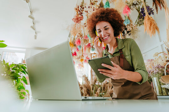 Smiling Afro florist talking on smart phone using tablet PC by laptop in flower shop