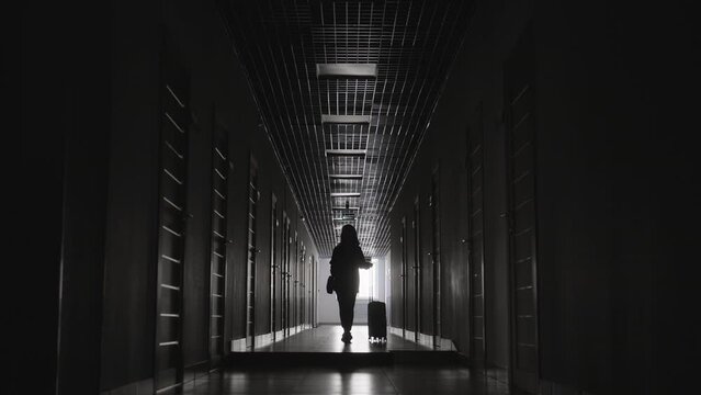 Full monochromatic shot of unrecognizable woman in dark corridor carrying suitcase walking out of hotel