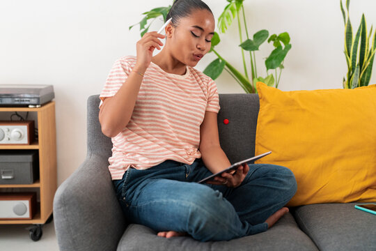 Young Asian female artist sitting on sofa with crossed legs wearing striped t shirt drawing sketch on graphic tablet with stylus during free time
