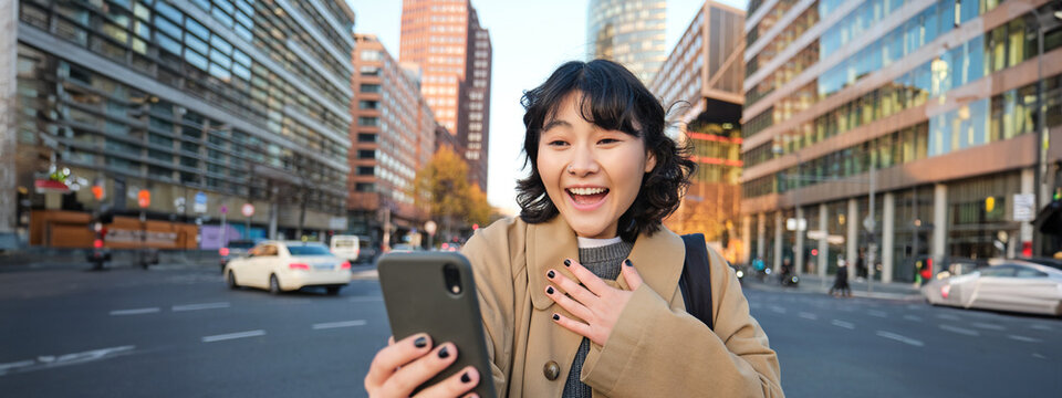 Image of korean girl video chats with smartphone, looks at her phone with surprised and amazed face, hears great news, stands on street