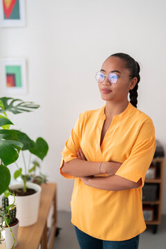 Portrait of self assured young female office worker wearing yellow shirt and glasses crossing arms while standing near potted plants in modern workspace