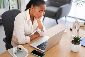 Young Asian female employee sitting at wooden desk and watching video on laptop during workday in contemporary workspace
