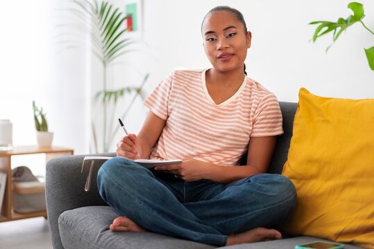 Young Asian female sitting on sofa wearing striped t shirt writing on notepad with pen during free time