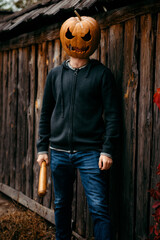 A man with a pumpkin on his head holds a bat in his hands and smiles ominously against the...