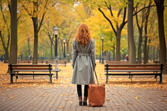 Rear view of young woman with coat and luggage in background of autumn park. Impressive concept of farewell and departure.