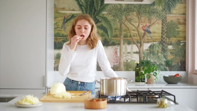Young caucasian woman cuts cabbage for making soup. Female stands in the kitchen at the stove and tastes the cabbage. Cooking in home kitchen with pleasure concept