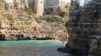  large cliff on the seashore, rocks by the sea, buildings built on the rocks right on the seashore, Polignano a Mare, rocky coast, turquoise water, people bathing in the sea