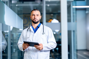 Handsome doctor with tablet computer gadget posing in hospital looking at us. Online medical consultation background with copy space.
