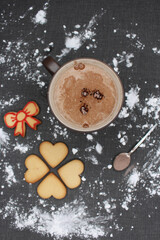 Black tablecloth full of flour, after having made homemade sweets for Christmas celebration. A mug of hot chocolate with lumps or coffee, heart gingerbread and lace cookies and a spoon with cocoa.