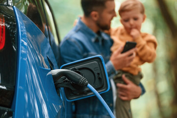 Close up view of wire in the socket. Father with his little son are waiting for electric car to charge - obrazy, fototapety, plakaty