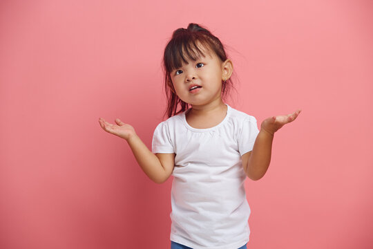 Confused little asian girl shrugs shoulders and expresses a question expression, says I don't know, has a difficult choice, standing isolated on pink background wearing in white t-shirt.