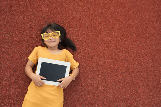 A little girl, 7-8 years old, wearing a yellow T-shirt and wearing yellow glasses, lies down and listens to music after exercising in the stadium. Girl playing sports happily